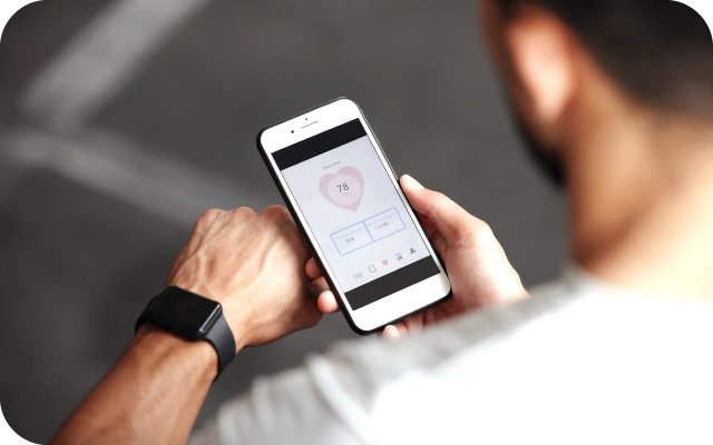 Vīrietis tur rokā mobilo telefonu, kurā redzama veselības lietotne ar sirds ritma rādījumu 78 sitieni minūtē, un vienlaikus skatās viedpulkstenī uz savas rokas.