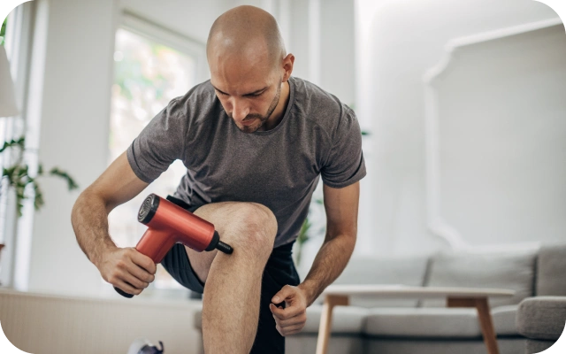 Vīrietis, notupies uz viena ceļa, mājas vidē ar masāžas pistoli masē kājas muskuļus. Fonā dīvāns un gaiša, ērta viesistaba.