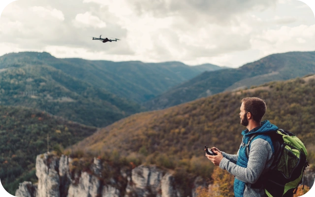 Vīrietis stāv kalnainā ainavā uz klints malas un ar tālvadības pulti vada debesīs lidojošu dronu. Fonā plaši, zaļi kalni un mākoņainas debesis.