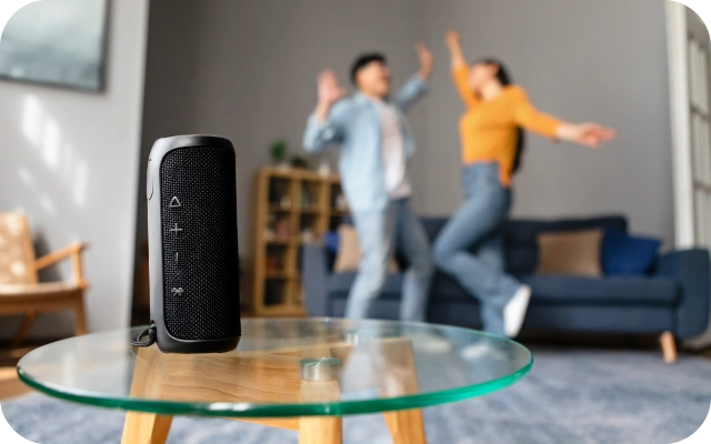 Uz stikla galdiņa priekšplānā novietots melns bezvadu skaļrunis. Fonā divi cilvēki dzīvojamajā istabā dejo.