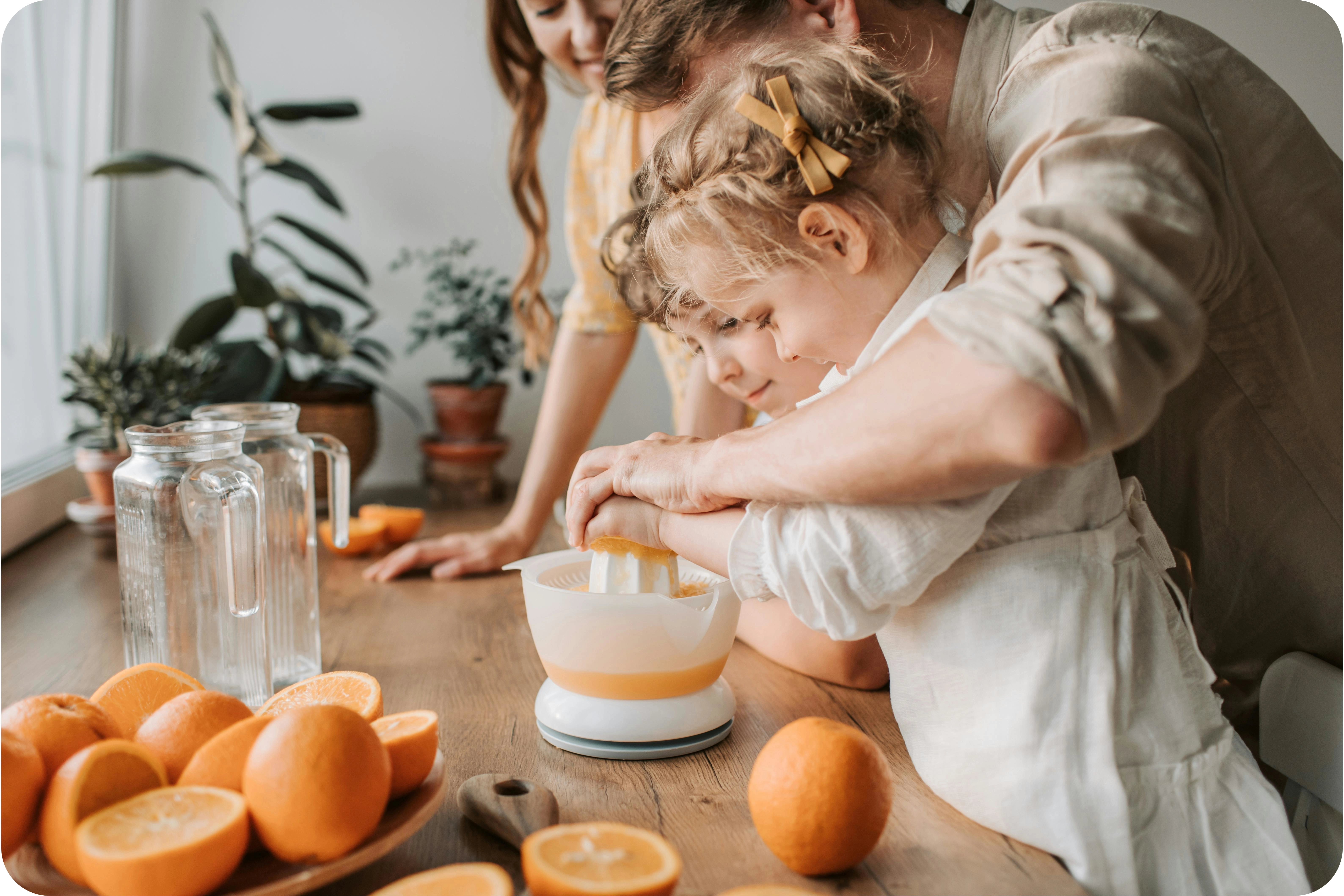 Ģimene kopīgi spiež apelsīnu sulu uz koka galda; uz galda ir apelsīni, stikla krūze un augiem podos.