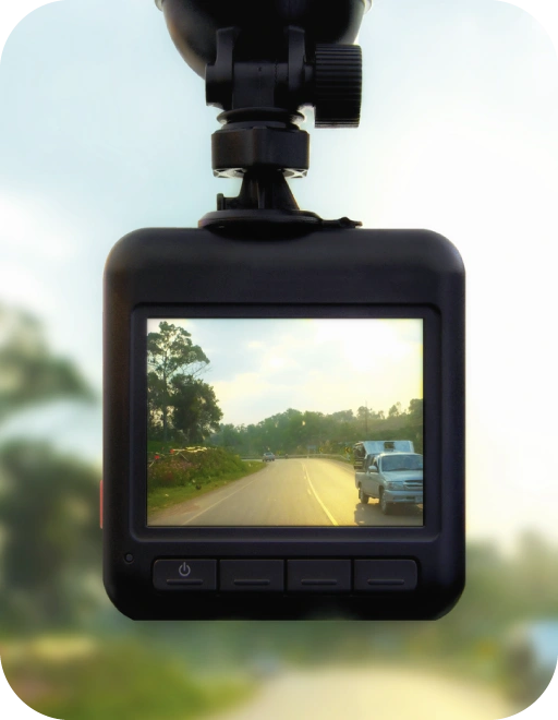 Automašīnas videoreģistrators ar ekrānu, kurā redzams dienas skats uz divvirzienu ceļu ar priekšā braucošu auto un kokiem ceļa malās.