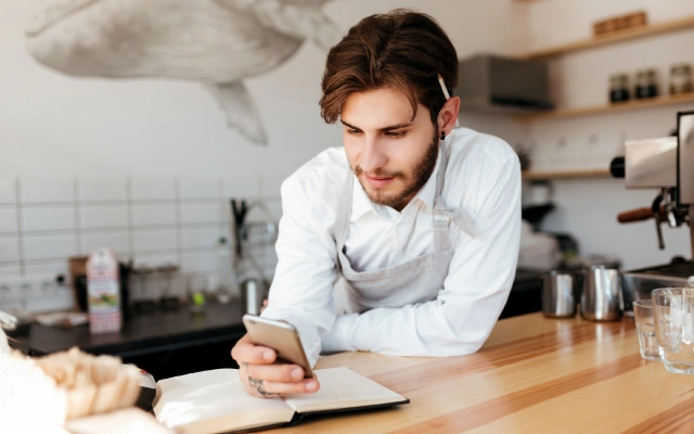 Vīrietis baltā kreklā, priekšautā ar zīmuli aizliktu aiz auss, stāv atspiedies uz koka letes, skatās mobilajā telefonā ko tur vienā rokā; uz letes atvērta piezīmju grāmatiņa, fonā pie sienas zīmējums ar vali un virtuves piederumi.