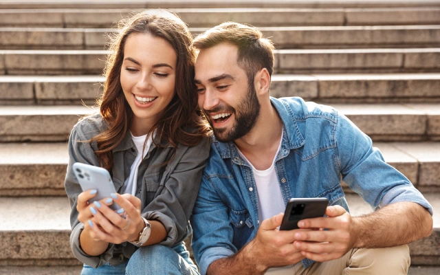 Sieviete un vīrietis ikdienišķā apģērbā sēž uz kāpnēm un skatās mobilajā telefonā.