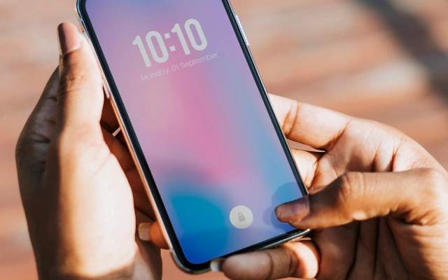 Mobilais telefons sievietes rokās; telefona ekrānā redzams laiks 10:10 un datums – pirmdiena, 1. septembris.