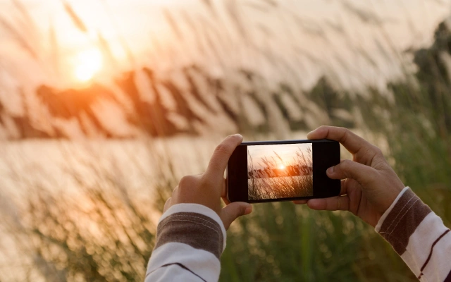 Cilvēks fotografē saulrietu pie ezera ar mobilo telefonu, fonā garas zāles stiebri un silta saules gaisma.