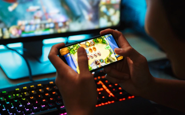 Cilvēks spēlē spēli mobilajā telefonā, sēžot pie datora ar izgaismotu klaviatūru un monitoru fonā.