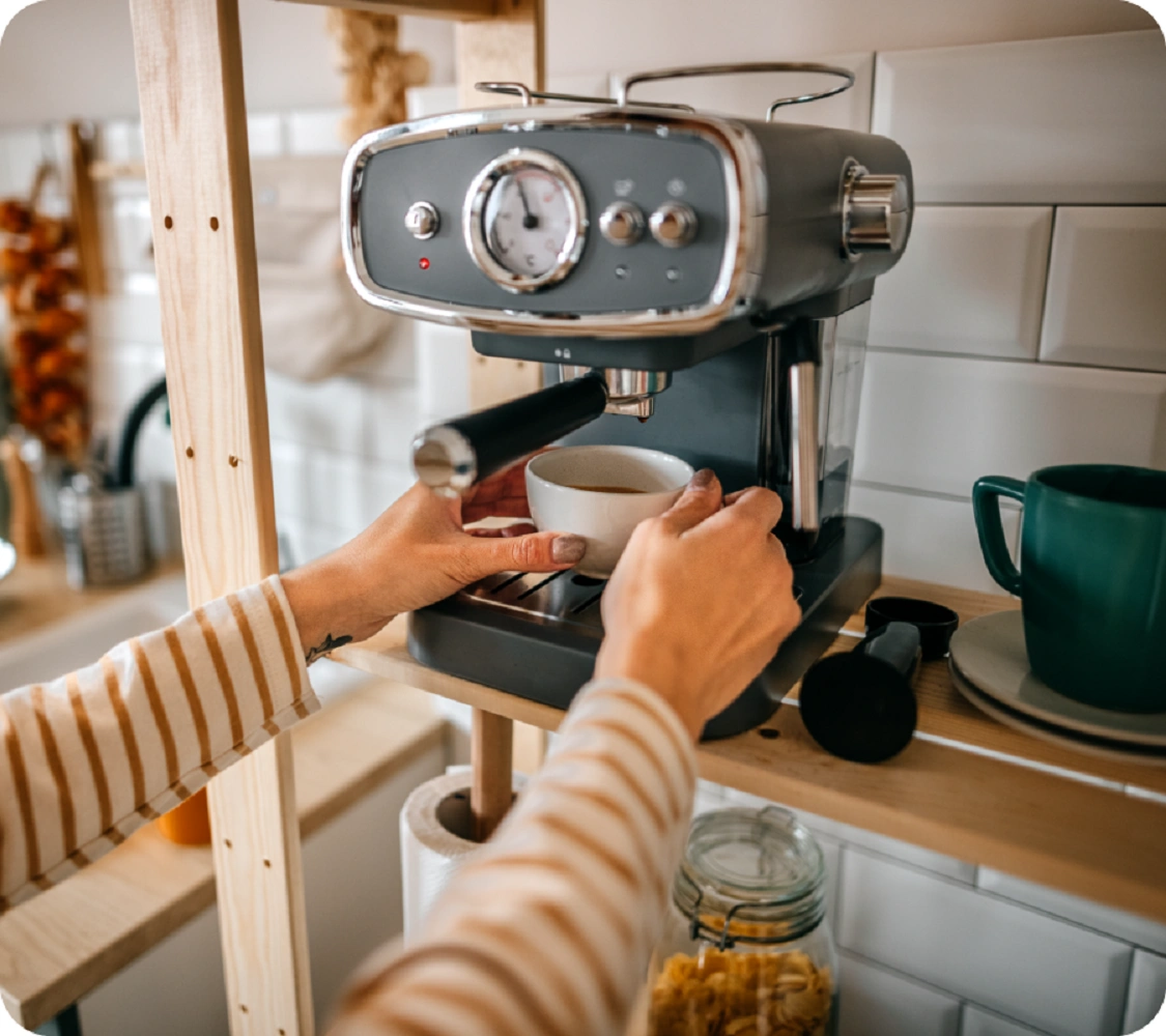 Cilvēks gatavo kafiju espresso kafijas automātā, turot baltu krūzi zem filtra sviras; fonā zaļa krūze, šķīvis un virtuves piederumi.