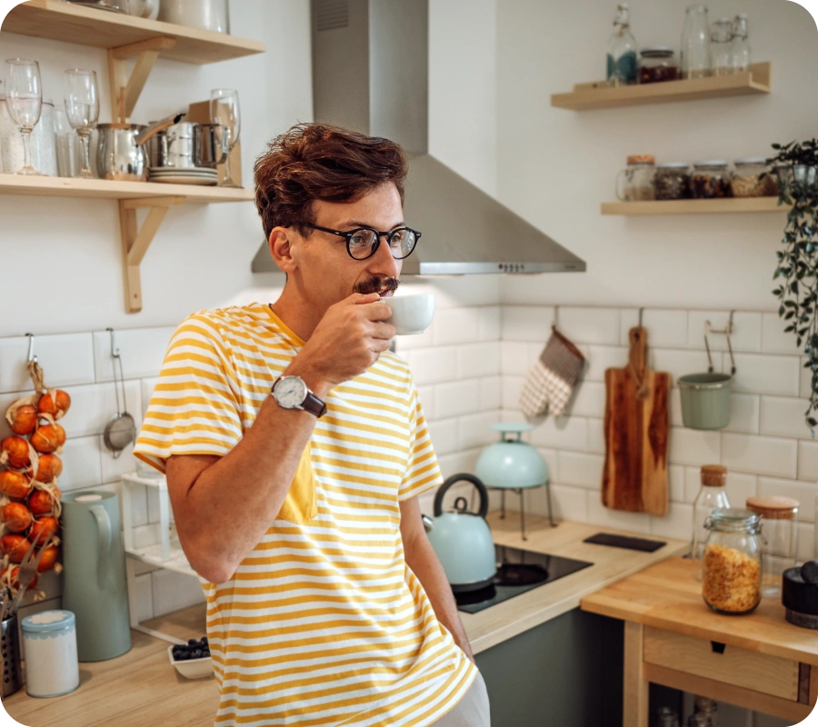 Vīrietis ar brillēm dzeltenā svītrainā T-kreklā dzer no baltas krūzes virtuvē ar koka plauktiem; fonā zila tējkanna uz plīts un virtuves piederumi uz galda.