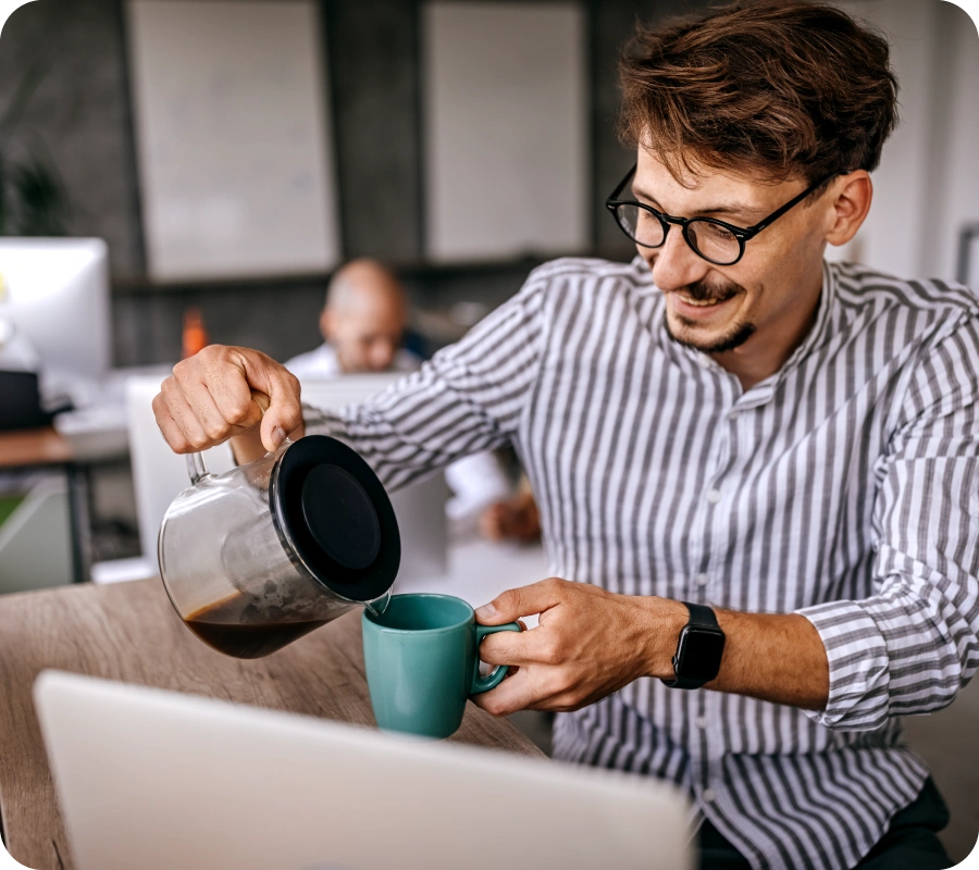 Vīrietis ar svītrainu kreklu lej kafiju zaļā krūzē no stikla kafija kannas, fonā personas, kas strādā pie datora.