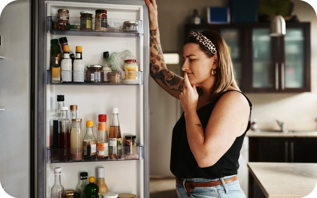 Sieviete stāv virtuvē pie atvērtām ledusskapja durvīm, skatoties ledusskapja iekšpusē; ledusskapja durvju plauktos novietotas dažādas mērces, burciņas un dzērienu pudeles.
