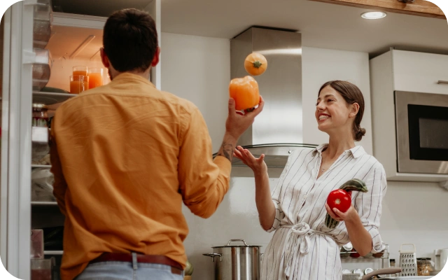Virtuvē sieviete un vīrietis stāv pie ledusskapja; vīrietis skatās iekšā atvērtajā ledusskapī turot rokās dzeltenu papriku, sieviete žonglē ar sīpolu, otrā rokā turot sarkanu tomātu un cukinī, fonā virtuves tehnika.