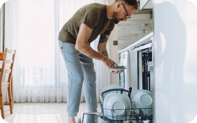 Vīrietis virtuvē izņem traukus no trauku mazgājamās mašīnas — tur rokās nelielu šķīvju kaudzīti un ir noliecies virs apakšējā trauku mazgājamās mašīnas groza.