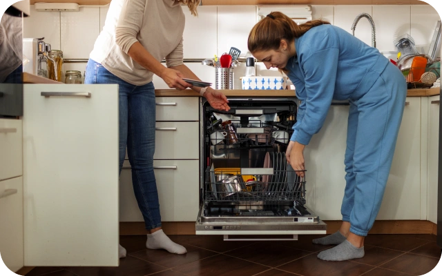 Divas sievietes virtuvē iztukšo trauku mazgājamo mašīnu — viena sieviete izņem traukus un padod tos otrai sievietei, kurai rokās jau ir daži šķīvji, fonā virtuves skapīši un dažādi virtuves piederumi.