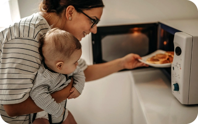 Sieviete tur bērnu vienā rokā, kamēr ar otru roku ievieto šķīvi ar ēdienu mikroviļņu krāsnī; fonā balta virtuve ar skapīšiem.