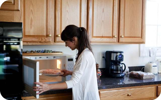 Sieviete baltā kreklā liek šķīvi ar ēdienu mikroviļņu krāsnī,fonā virtuvē ar koka skapīšiem, virtuves piederumi — kafijas automāts un blenderis.