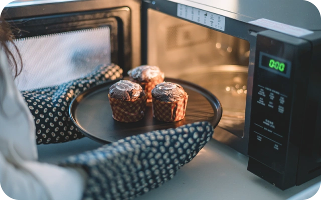 Persona ar virtuves cimdiem rokās izņem no mikroviļņu krāsns šķīvi ar trim cepumiem, kas apkaisīti ar pūdercukuru.