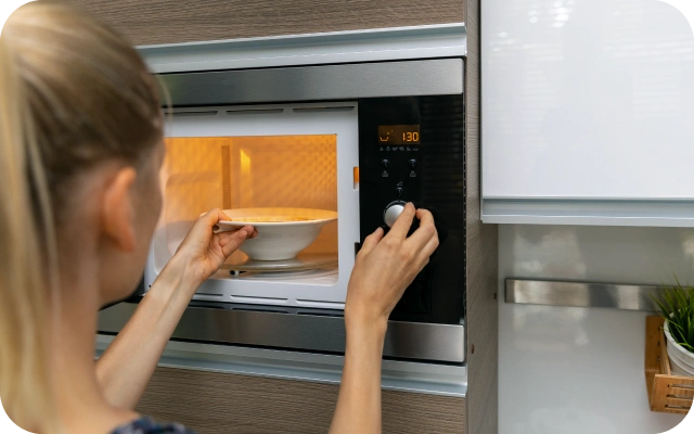 Sieviete ieliek bļodu ar ēdienu iebūvētā mikroviļņu krāsnī, iestatot taimeri uz 1:30.