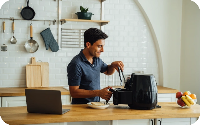 Vīrietis virtuvē izmanto aerogrilu — ar knaiblēm izņem ēdienu; fonā gaiša virtuve ar balta flīžu siena, pie sienas virtuves piederumi, virtuves skapīši ar koka virsmu.