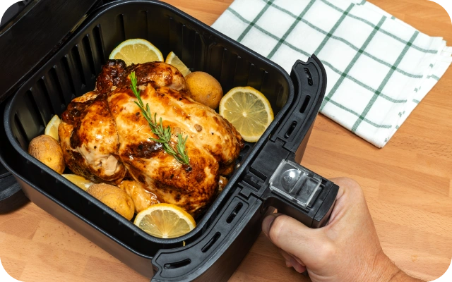 Kraukšķīgi brūni izcepta vista ar rozmarīna zariņu, citronu šķēlītēm un kartupelīšiem atrodas aerogrila grozā, kuru tur ar roku virs koka virsmas; fonā zaļš rūtainais dvielis.