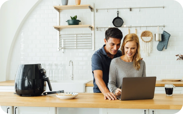 Vīrietis un sieviete virtuvē stāv pie koka darba virsmas viens otram blakus un skatās portatīvajā datorā; uz letes novietots melns aerogrils, šķīvis ar ēdienu un glāze ar tumšu dzērienu; fonā balta flīžu siena un plaukti ar virtuves priekšmetiem.