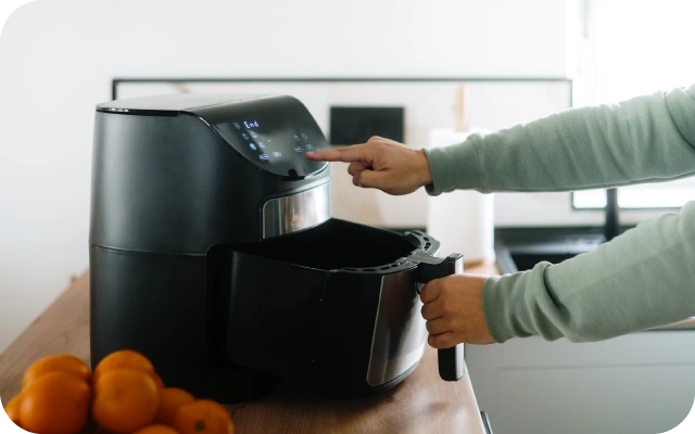 Persona virtuvē lieto melnu aerogrilu — ar vienu roku tur aerogrila grozu, ar otru piespiež pogu uz ierīces digitālā paneļa; blakus uz koka virsmas ir bļoda ar apelsīniem.