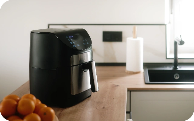 Virtuves darba virsma ar melnu aero ar digitālo displeju, blakus bļoda ar apelsīniem, labajā pusē papīra dvieļu turētājs pie melnas izlietnes.