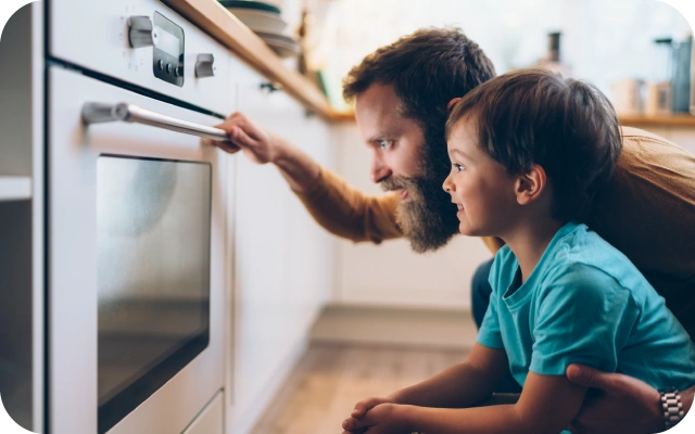 Vīrietis un bērns notupušies pie cepeškrāsns vēro kā gatavojas ēdiens; vīrietis vienu roku uzlicis uz cepeškrāsns roktura, bet ar otru roku apķēris bērnu, fonā virtuves skapīši.