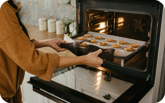 Persona brūnā džemperī ar krāsns cimdiem izņem karstu paplāti ar svaigi izceptiem cepumiem; cepumi novietoti rindās uz cepamā papīra, krāsns iekšpuse izgaismota, fonā balti trauki un augs uz darba virsmas.