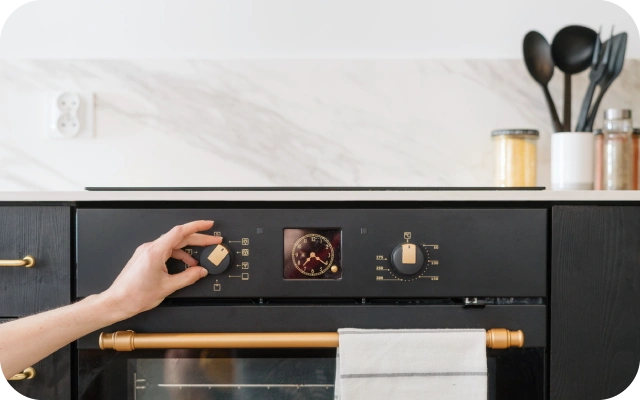 Roka pagriež vienu no cepeškrāsns pogām uz melna vadības paneļa; fonā marmora raksta flīžu siena, virtuves piederumi un balts dvielis uz krāsns roktura.