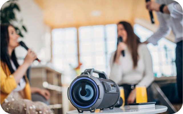 Trīs cilvēki dzied karaoke ar mikrofoniem rokās, priekšplānā uz galda novietots bezvadu skaļrunis.