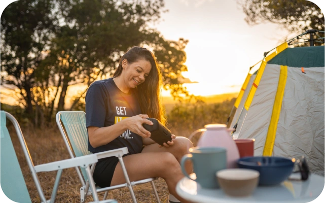Sieviete sēž saliekamā krēsla pie telts, turot rokās bezvada skaļruni; priekšplānā galds ar krūzēm un bļodām, fonā koki un zeltains saulriets.