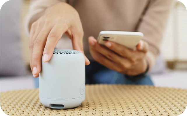 Sieviete tur rokā baltu mobilo telefonu un ar otru roku pieskaras nelielam cilindriskam bezvadu skaļrunim, kas novietots uz pīta galda.