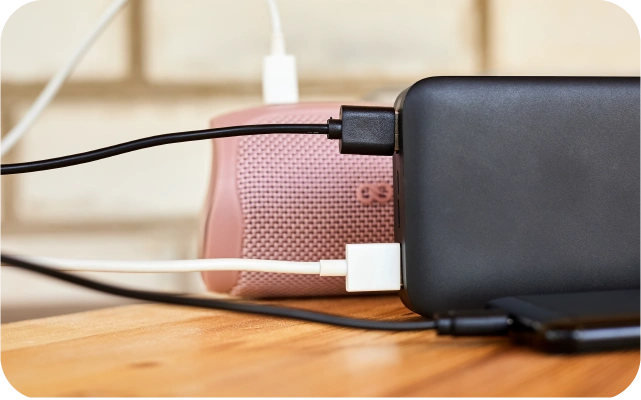 Melns portatīvais lādētājs uz koka virsmas ar pieslēgtiem USB kabeļiem- vienu, kas lādē rozā Bluetooth skaļruni, otru, kas lādē melnu mobilo telefonu.