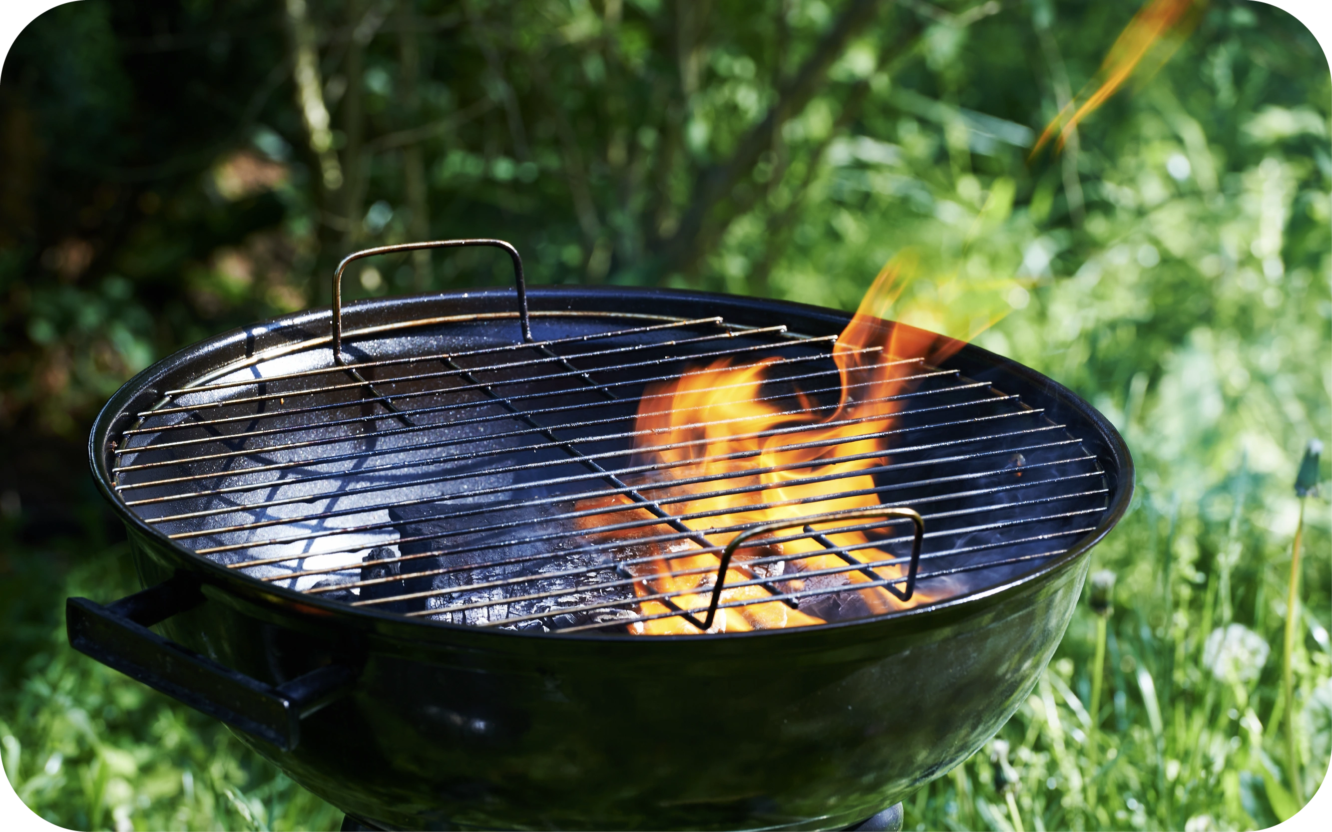 Melns, apaļš grils ar metāla restēm un uguns liesmu, fonā zāliens un krūmi.
