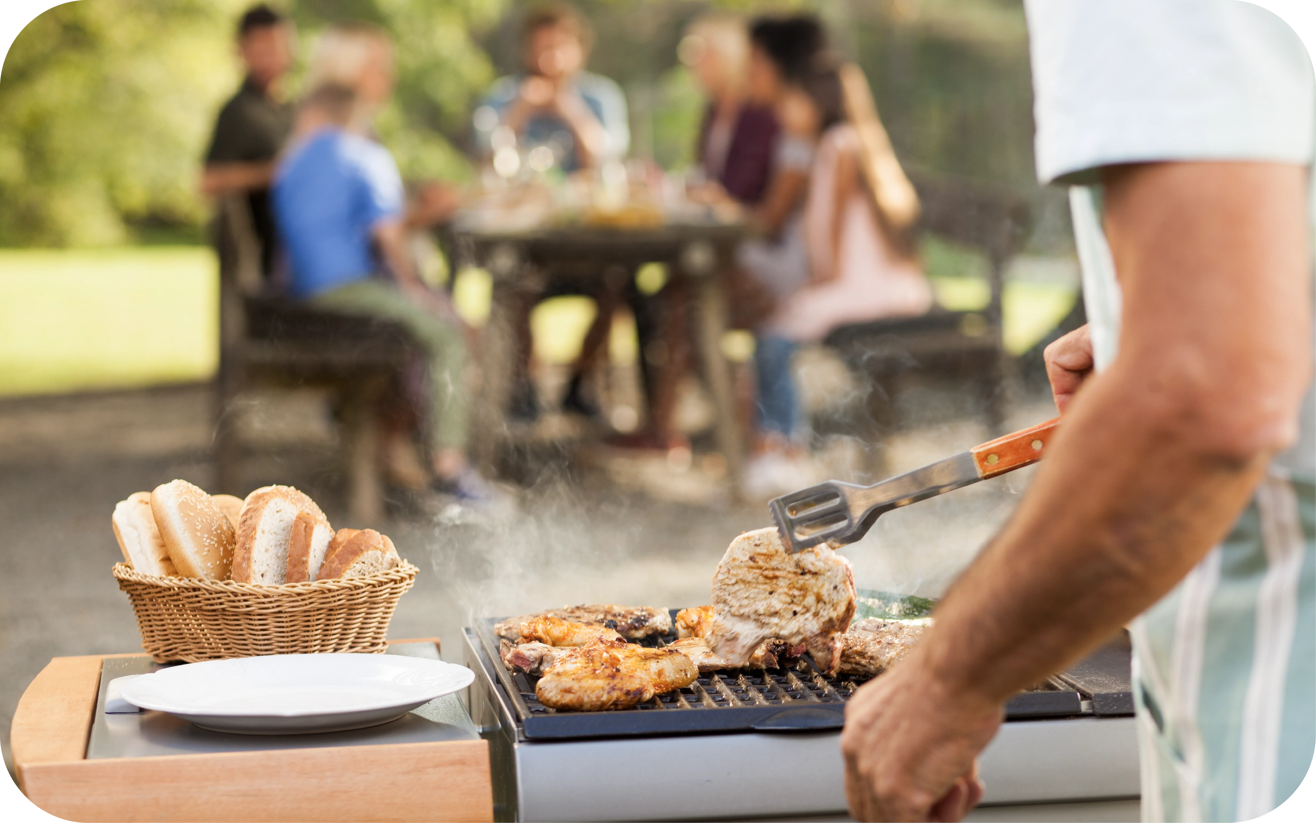 Cilvēks grilē gaļu, blakus uz grila plauktiņa ir grozs ar maizi un šķīvis, fonā pie galda sēž un sarunājas cilvēki.