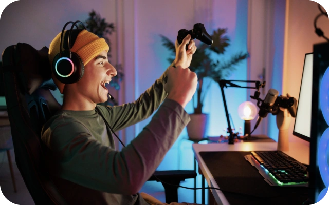 Jaunietis ar austiņām un mikrofonu spēlē videospēli pie datora galda, turot rokās spēļu kontrolieri; uz galda ir monitors, klaviatūra un pele.