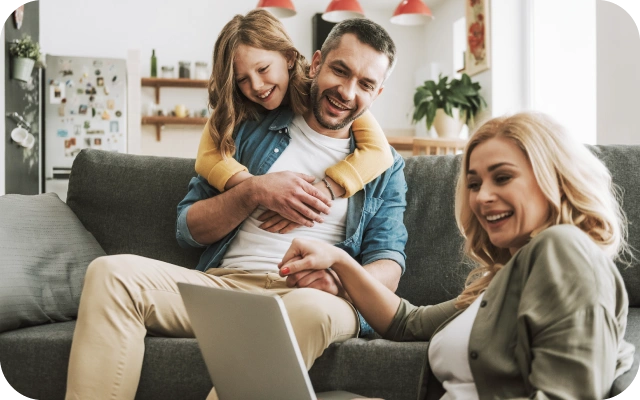 Ģimene sēž uz dīvāna mājīgā viesistabā — bērns ir apķēris vīrieti no mugurpuses, sieviete klēpī tur portatīvo datoru.