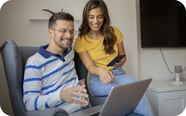 Sieviete un vīrietis sēž uz dīvāna — viens tur portatīvo datoru, otrs mobilo telefonu.