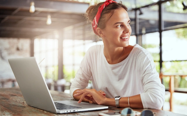 Sieviete ar sarkanu galvas lenti un baltu kreklu sēž pie galda ar portatīvo datoru, viedpulksteni ap roku un saulesbrillēm uz galda; fonā stiklota siena.