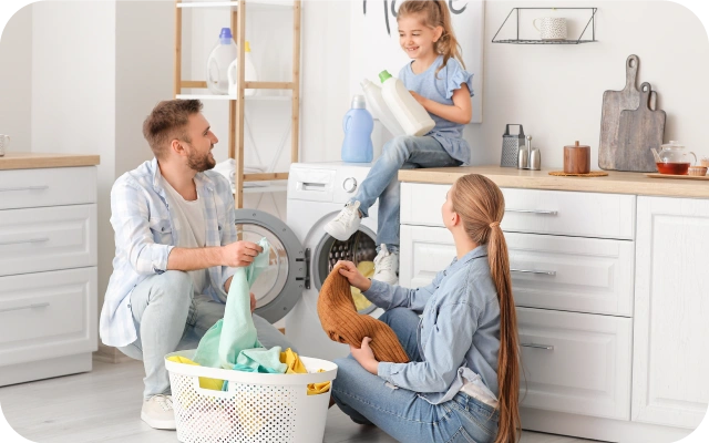 Vīrietis un sieviete tup pie atvērtas veļas mašīnas ar veļu grozu pie kājām un apģērba gabaliem rokās, bērns sēž uz galda virsmas virs veļas mašīnas un tur rokās tīrīšanas līdzekļus; fonā skapīši un plauki.