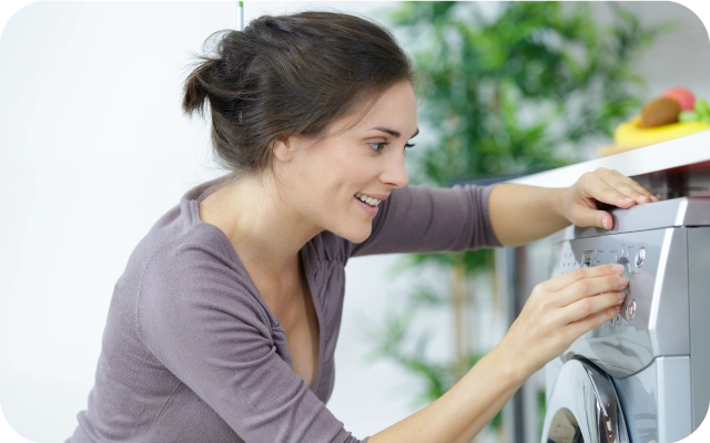 Sieviete iestata veļas mazgāšanas režīmu, nospiežot pogas uz veļas mašīnas vadības paneļa; fonā puķe un augļu bļoda.