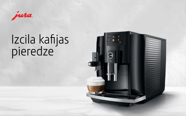 Melns automātiskais kafijas automāts (JURA E8 Piano) ar piena putotāju gatavo kapučīno stikla krūzē; balts fons.